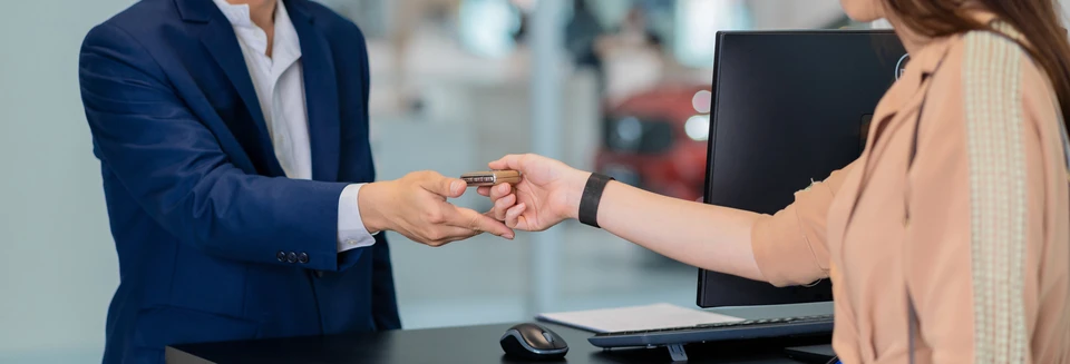 Conseiller automobile en tenue professionnelle présentant les caractéristiques d'un véhicule d'occasion à un client debout face à la voiture, dans l'espace de parc du garage. Le conseiller gestualise vers différentes zones du véhicule tandis que le client l'écoute attentivement. Environnement de garage automobile indépendant, lumière naturelle, atmosphère de confiance et professionnalisme.