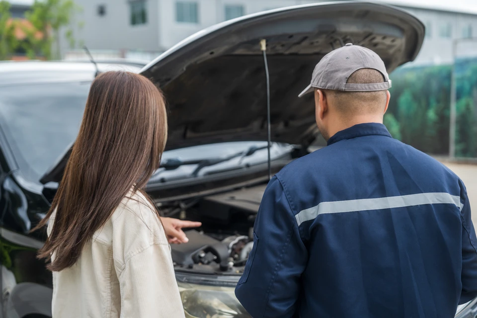 Atelier automobile moderne avec mécanicien en combinaison bleue consultant documents jaunes avec cliente en chemisier beige près d'un véhicule noir au capot ouvert
